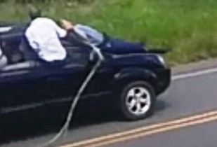 Segurança amarrado no capô e um bandido fortemente armado ao seu lado com o carro em alta velocidade.